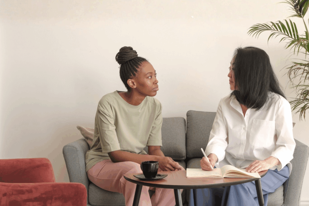 Two professional women having a supportive mentoring conversation on a sofa, one listening and one writing notes.